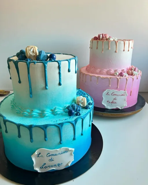 Due drip cake per Comunione su basi separate, una azzurra e una rosa, decorate con macarons, perle zuccherate e targhette con i nomi dei festeggiati. La foto mette in evidenza il contrasto cromatico e l'impostazione scenografica delle torte.
