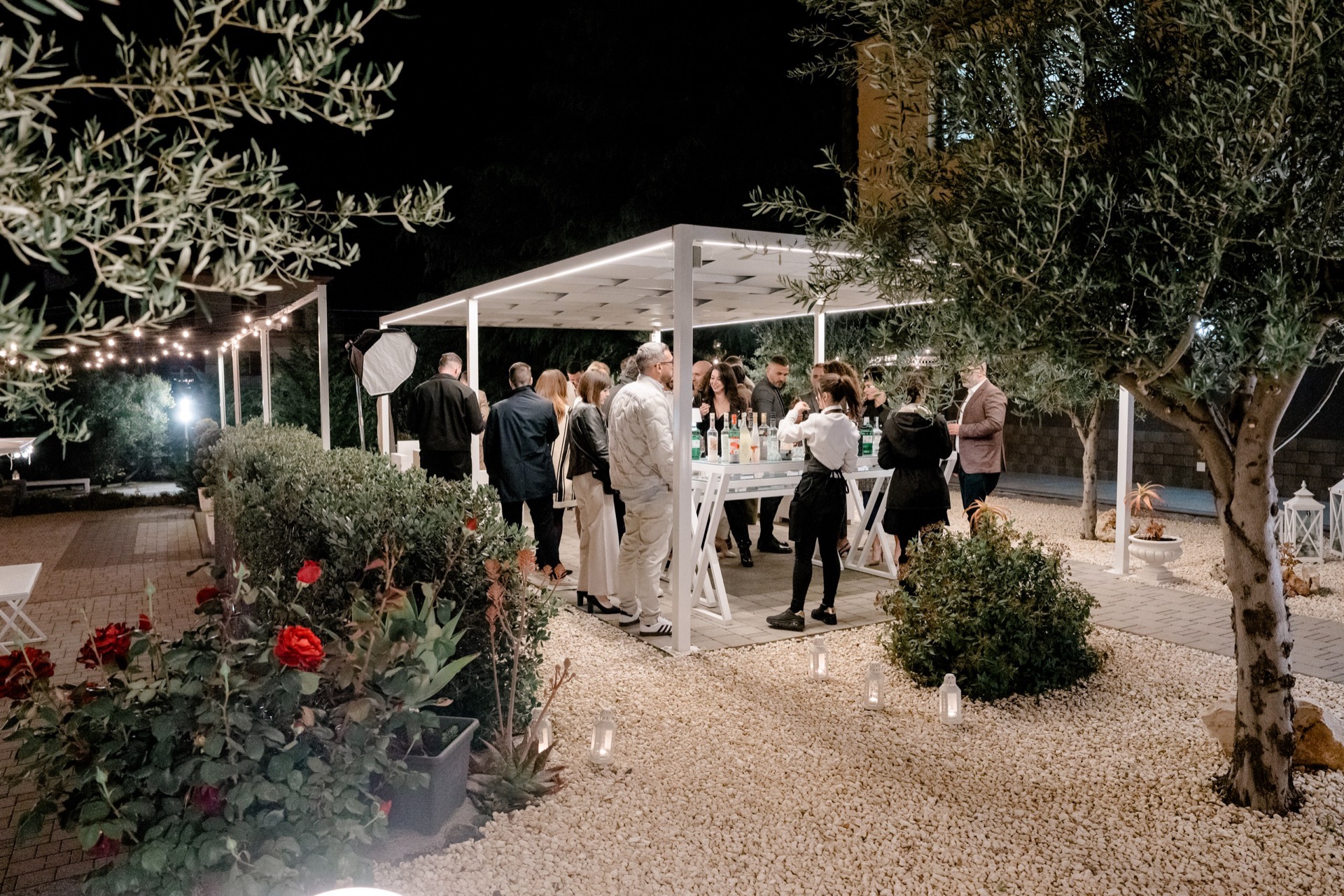 Angolo relax e bar sotto una pagoda moderna, con ospiti raccolti attorno al banco drink e il giardino illuminato da luci calde. La ghiaia bianca, gli alberi e le rose in primo piano rendono la scena elegante e notturna.