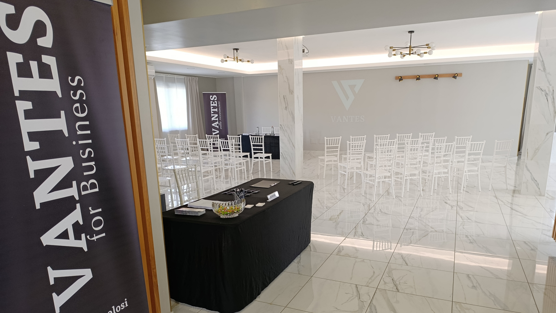 Sala per eventi aziendali con sedie disposte in file, tavolo relatori e banner VANTES visibili all'ingresso e sullo sfondo. L'ambiente è minimale, luminoso e adatto a presentazioni o meeting.