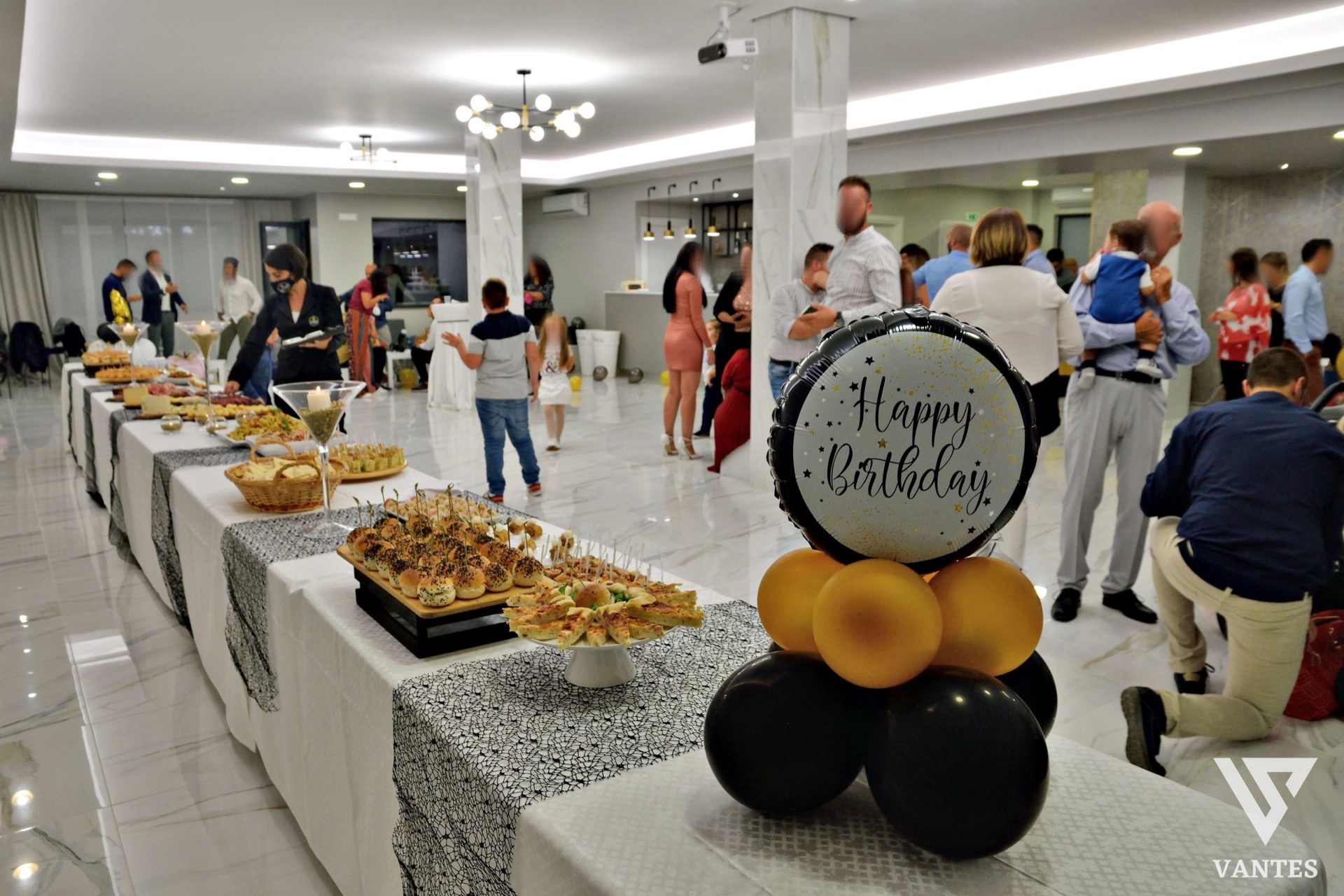 Festa privata in sala con lungo buffet di finger food, pallone decorativo 'Happy Birthday' e ospiti che conversano vicino alla zona buffet. L'ambiente è moderno, luminoso e organizzato per un compleanno o evento familiare.