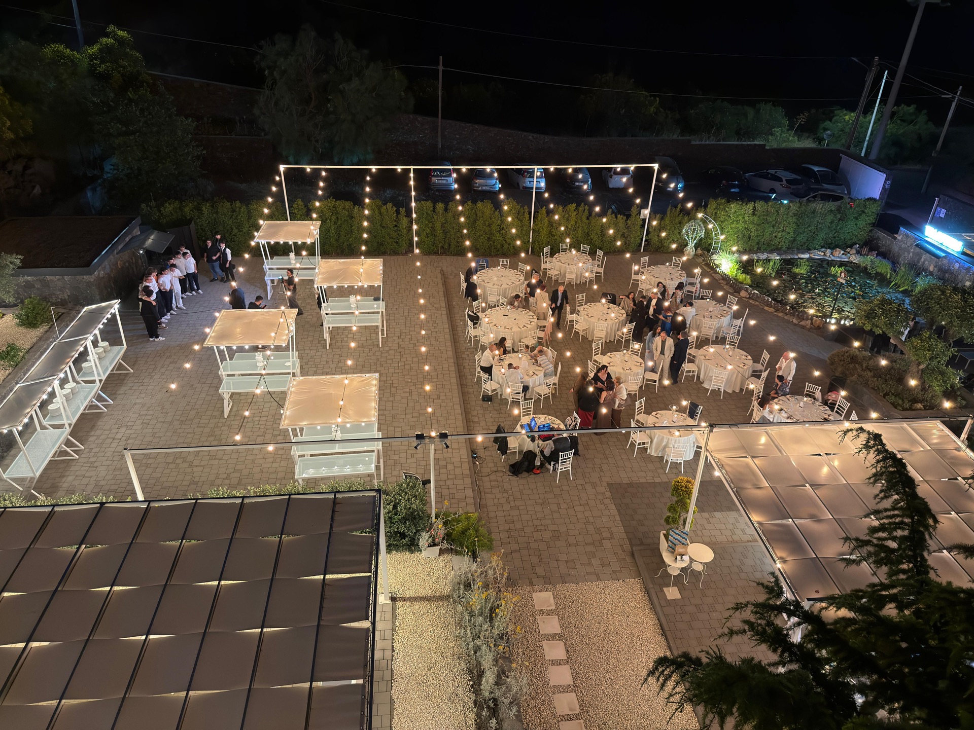 Vista aerea notturna della location con tavoli rotondi, buffet e aree relax distribuite nel cortile. Le luci sospese e il laghetto illuminato evidenziano la geometria dell'allestimento dall'alto.