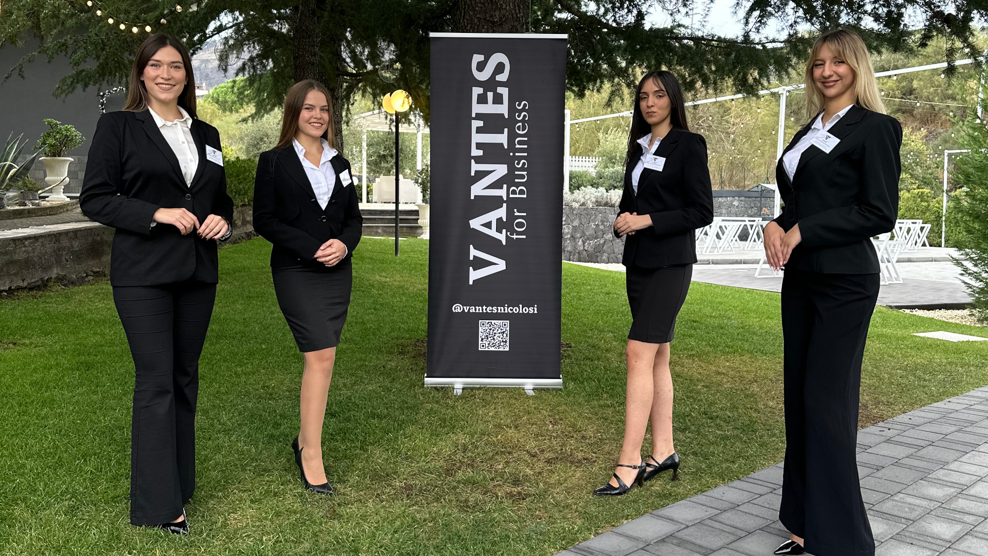 Quattro persone dello staff in divisa elegante posano sul prato accanto a un banner Vantes for Business. Sullo sfondo si vedono il giardino della location, luci sospese e l'area esterna allestita per eventi aziendali.
