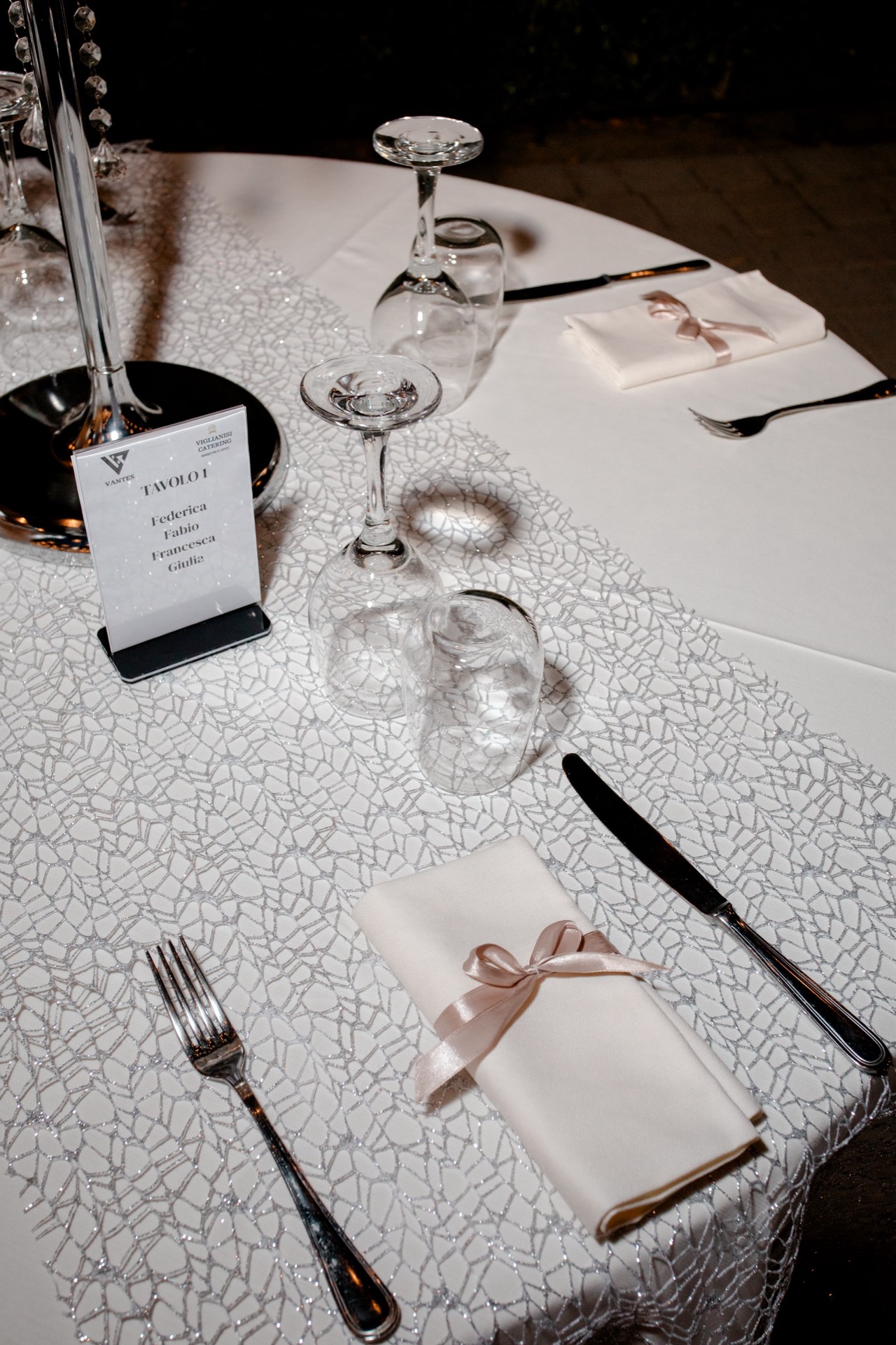 Dettaglio ravvicinato di una mise en place serale con runner chiaro lavorato, segnatavolo nominativo, calici capovolti e tovaglioli bianchi chiusi da un nastro rosa cipria. L'allestimento è elegante e ordinato, pensato per una cena all'aperto.