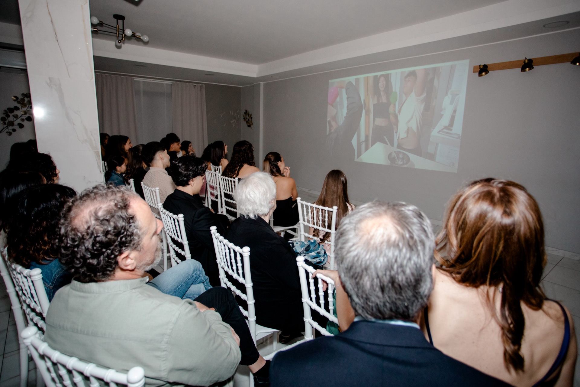 Ospiti seduti in sala durante la proiezione di un video ricordo su una grande parete bianca. La scena riprende il pubblico rivolto verso lo schermo in un momento raccolto della festa.