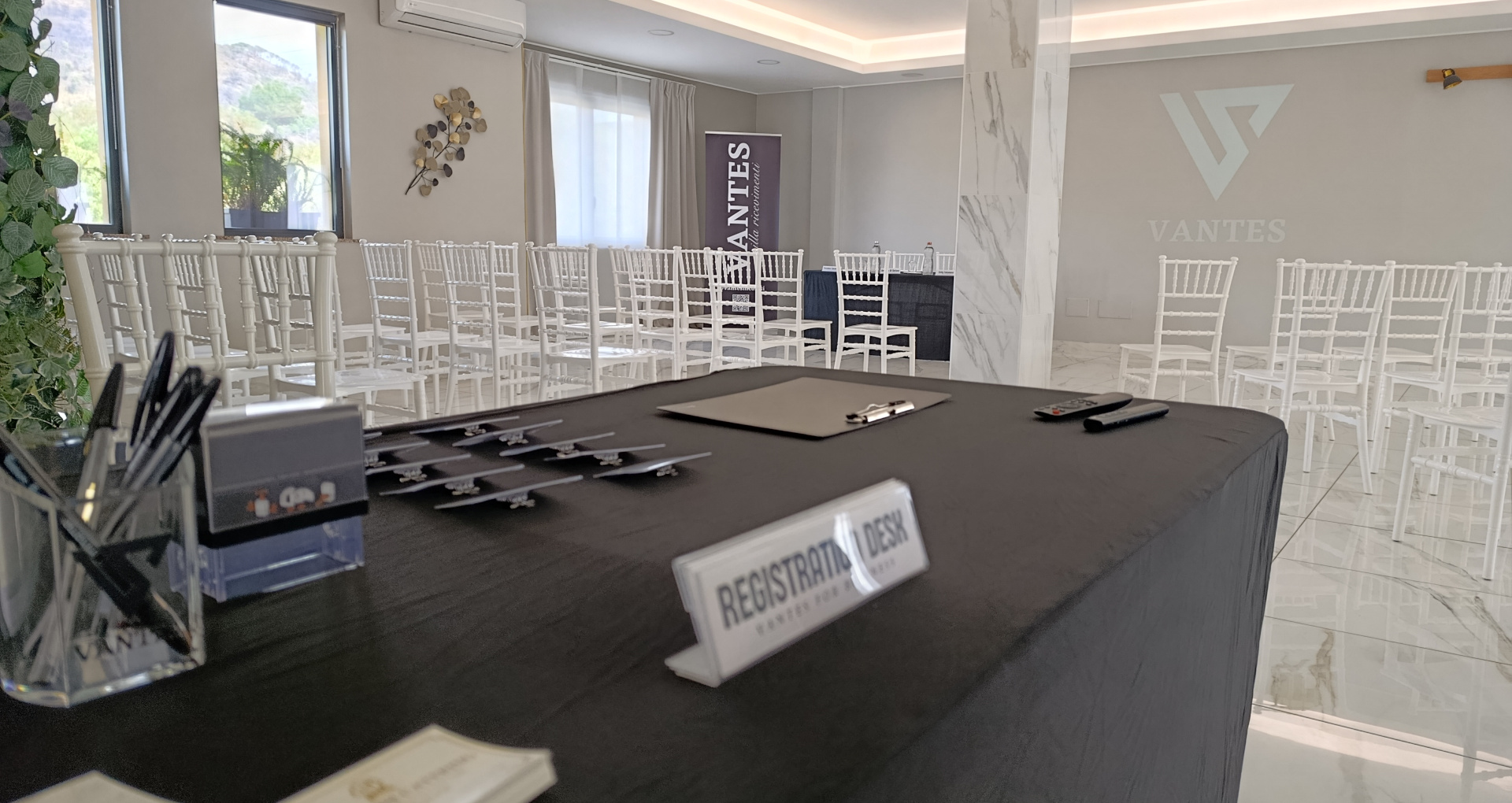 Desk registrazione per meeting allestito con penne, badge e cartello registration desk in primo piano, davanti a una sala con file di sedie bianche. Sullo sfondo si vedono il logo Vantes proiettato e un banner dedicato agli eventi business.