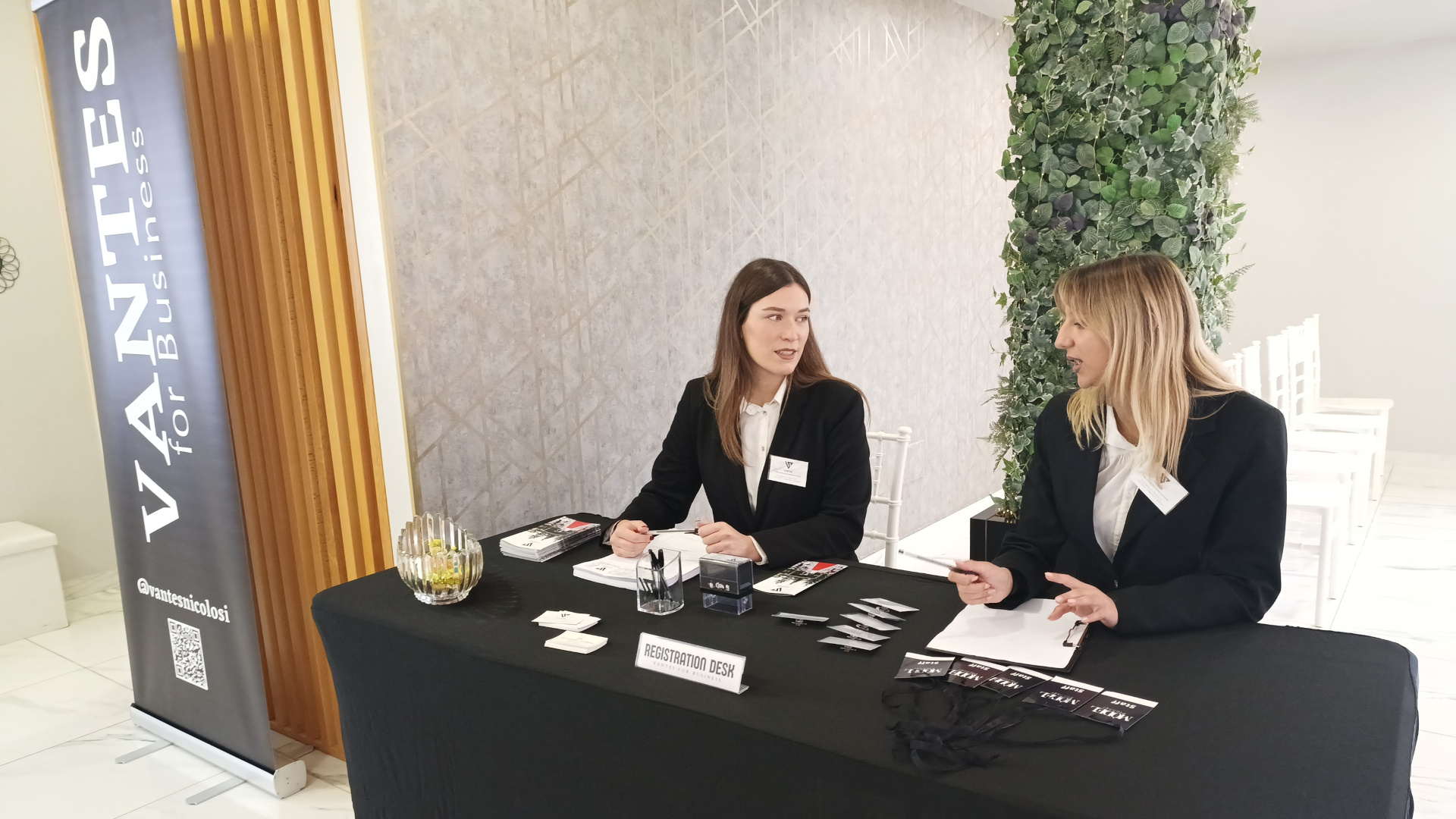 Desk registrazione presidiato da due persone dello staff in divisa, con badge, blocchi appunti e materiale informativo per un evento business. Accanto al tavolo si vede il banner Vantes for Business e sullo sfondo l'ingresso della sala con le sedie già disposte.