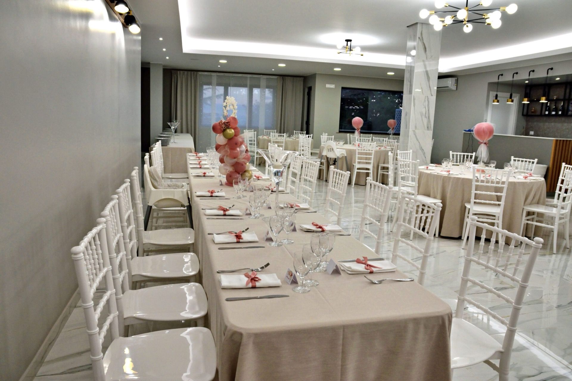Sala interna per battesimo con tavolata lunga, tavoli rotondi laterali e allestimento nei toni rosa cipria. I palloncini decorativi e la mise en place coordinata rendono l'ambiente delicato e festoso.