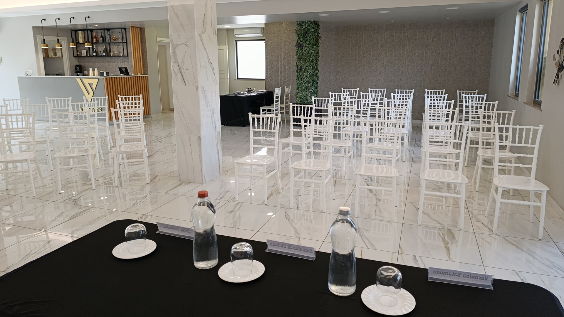 Sala conferenze allestita con file di sedie bianche, tavolo relatori in primo piano e banco bar sul fondo. L'ambiente è luminoso e ordinato, adatto a meeting, presentazioni e piccoli eventi professionali.