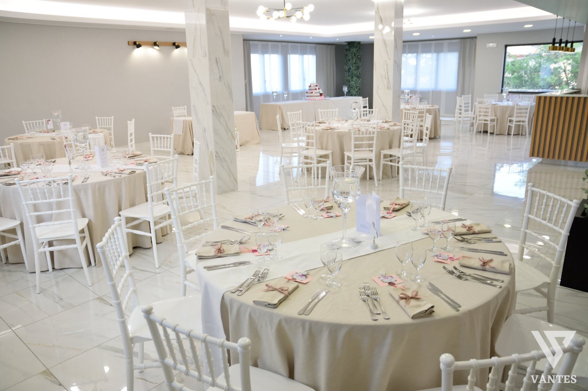 Sala per ricevimento elegante con tavoli rotondi apparecchiati, dettagli rosa cipria e tavolo della torta sul fondo. L'ambiente è luminoso, raffinato e adatto a matrimonio o cerimonia formale.