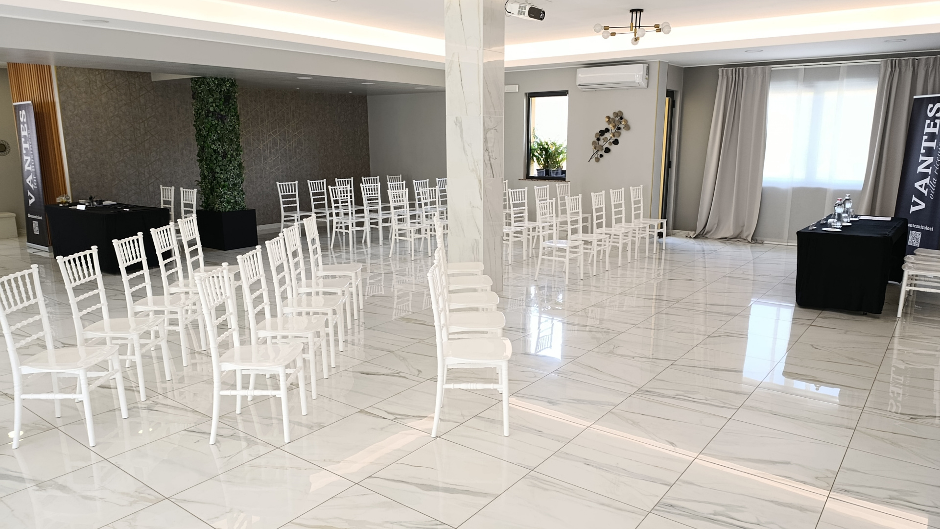 Sala meeting predisposta con file di sedute bianche, tavoli tecnici laterali e banner Vantes ai lati dell'ambiente. Lo spazio è ampio, luminoso e configurato per presentazioni o incontri professionali.