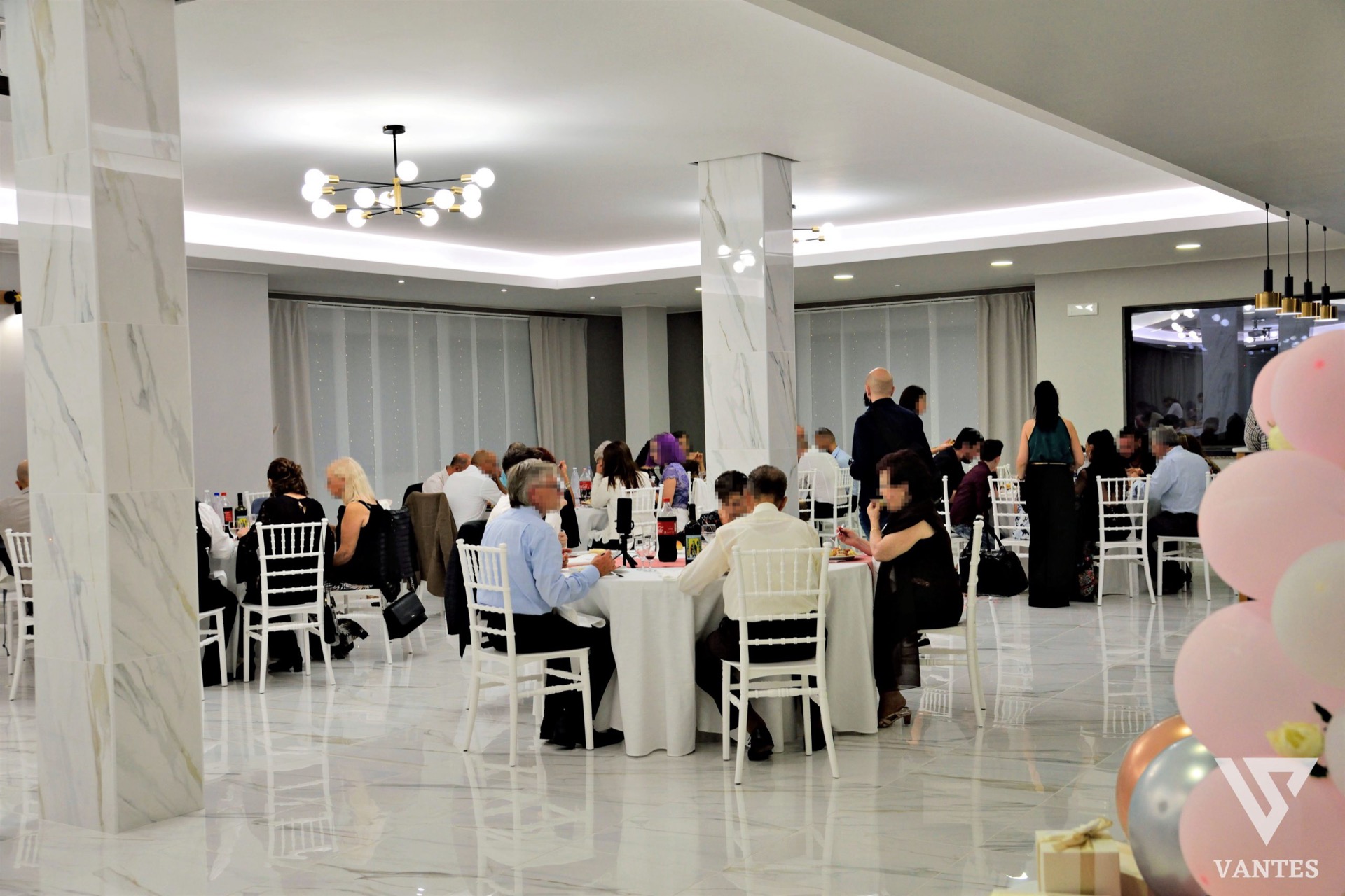 Sala ricevimenti durante una cerimonia con ospiti seduti ai tavoli rotondi e allestimento festa visibile sul lato dell'inquadratura. L'ambiente è elegante, luminoso e adatto a celebrazioni private e familiari.