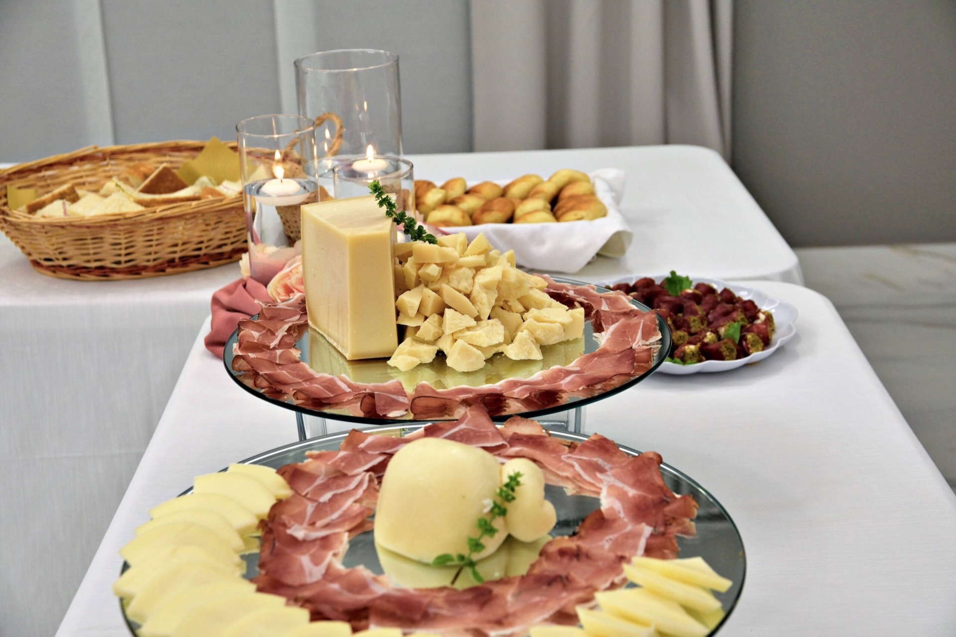 Buffet di salumi e formaggi allestito su alzate con prosciutto crudo, grandi forme di formaggio e cestino di pane sullo sfondo. La presentazione è ordinata ed elegante, pensata per un aperitivo o antipasto di cerimonia.
