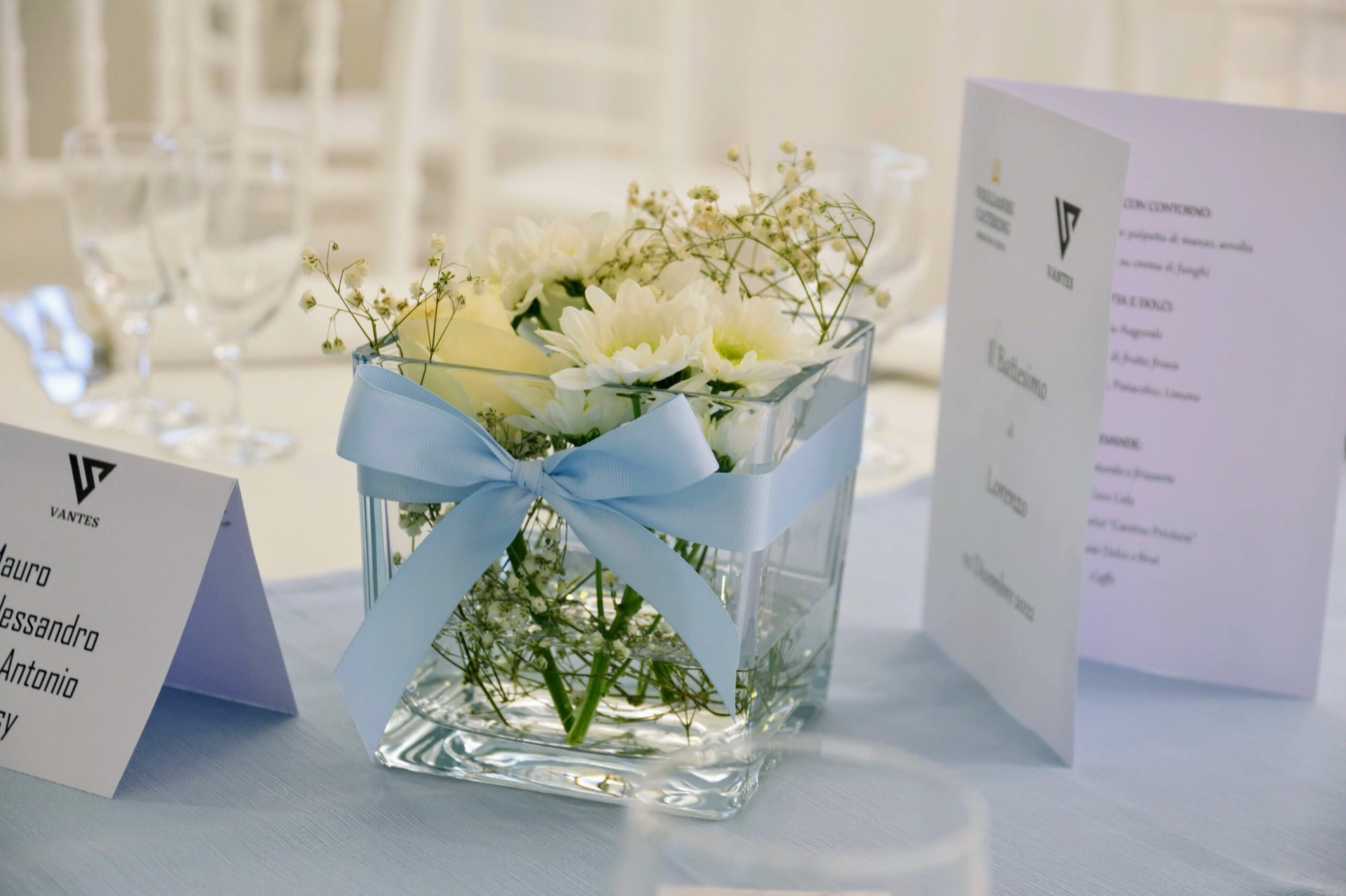 Dettaglio di tavolo da battesimo con centrotavola di fiori bianchi in vaso trasparente, nastro azzurro e menù o segnaposto coordinati. L'allestimento è luminoso, delicato e curato nei particolari della mise en place.
