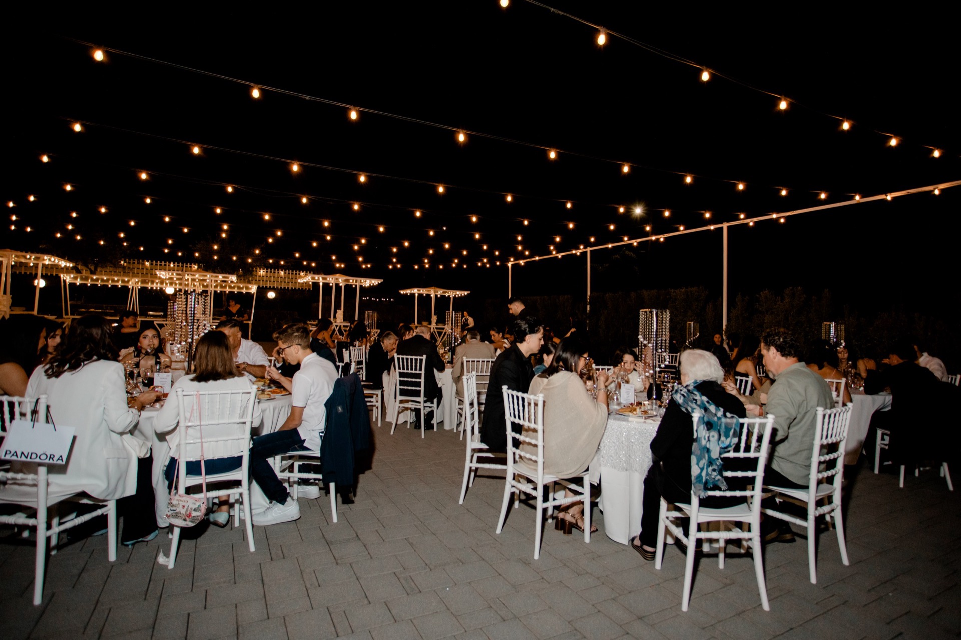 Ricevimento catering all'aperto con ospiti seduti a tavoli rotondi sotto una rete di luci sospese. La scena mostra una cena serale elegante e conviviale in uno spazio esterno ampio e curato.