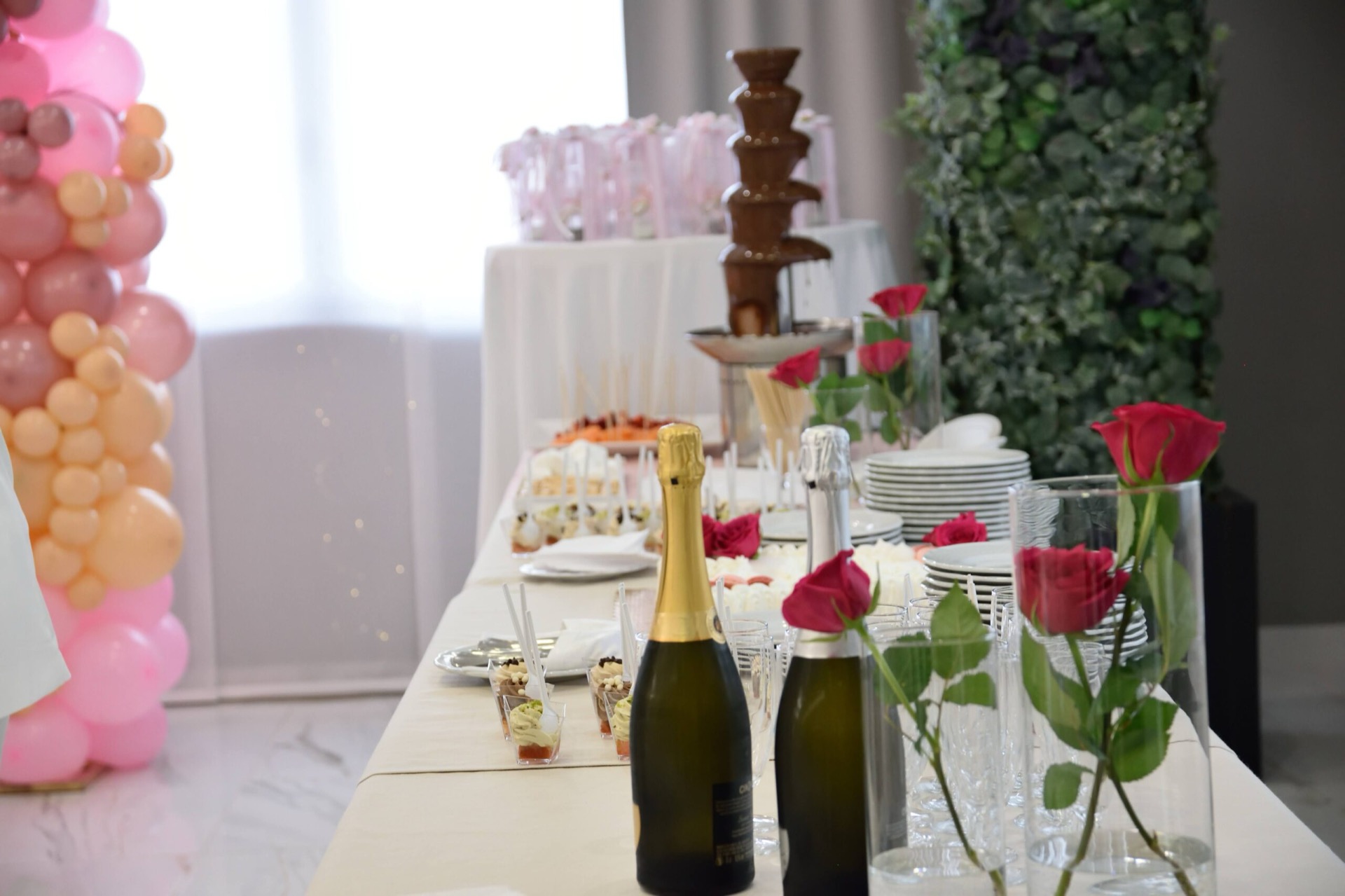 Tavolo dolci allestito per una cerimonia con dessert monoporzione, bottiglie, rose rosse in vaso e fontana di cioccolato sullo sfondo. Una colonna di palloncini rosa completa l'allestimento festoso della sala.