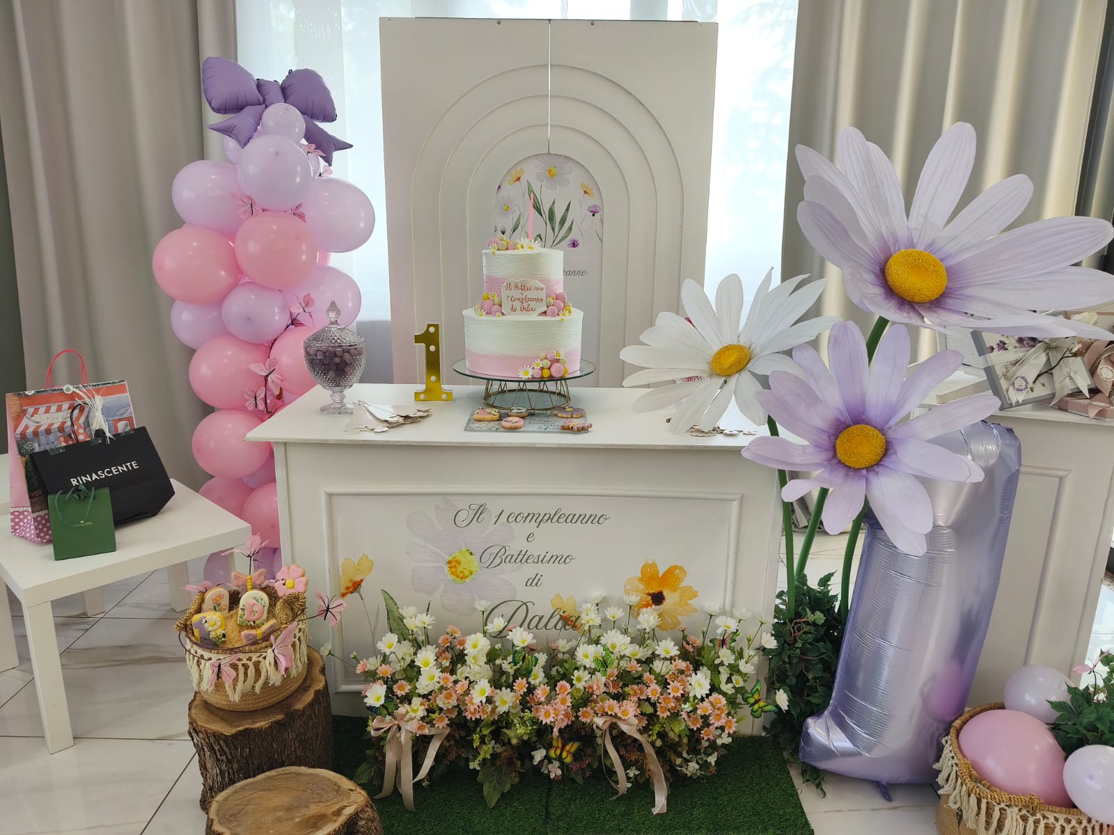 Allestimento in sala per primo compleanno e battesimo con torta centrale, grandi margherite decorative, colonna di palloncini rosa e numero 1 lilla. La scenografia è dolce e floreale, pensata come fondale principale della festa.