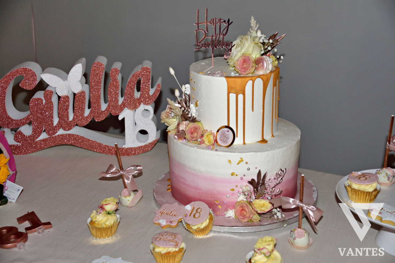 Torta a due piani per i 18 anni di Giulia, decorata con drip oro rosa, fiori pastello e piccoli dettagli coordinati. Sul tavolo si vedono anche cupcake e dolcetti abbinati all'allestimento della festa.